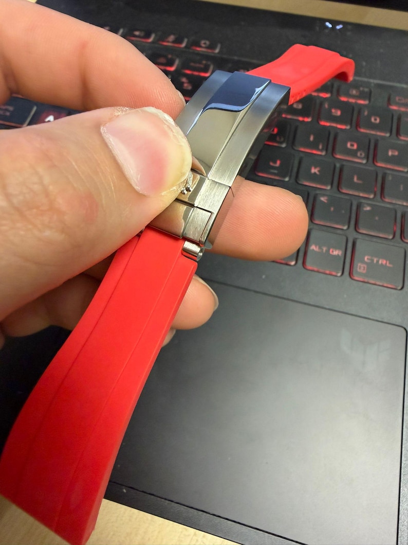 Peut inclure: Un bracelet de montre rouge avec un fermoir en acier inoxydable. Le bracelet est fait d'un mat&eacute;riau souple et est tenu par une main. Le fermoir est argent&eacute; et a une finition polie. L'arri&egrave;re-plan est un ordinateur portable noir.