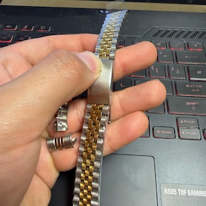 May include: Two-tone watch strap with silver and gold links. The strap is partially disassembled, with a clasp and several detached links visible. The watch strap is held against a laptop keyboard.