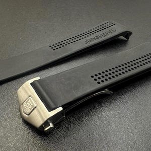 Pu&ograve; includere: Cinturino nero per orologio con il marchio "TAG Heuer" in rilievo. Il cinturino presenta una serie di piccoli fori e una chiusura color argento. Il cinturino &egrave; realizzato in materiale flessibile.