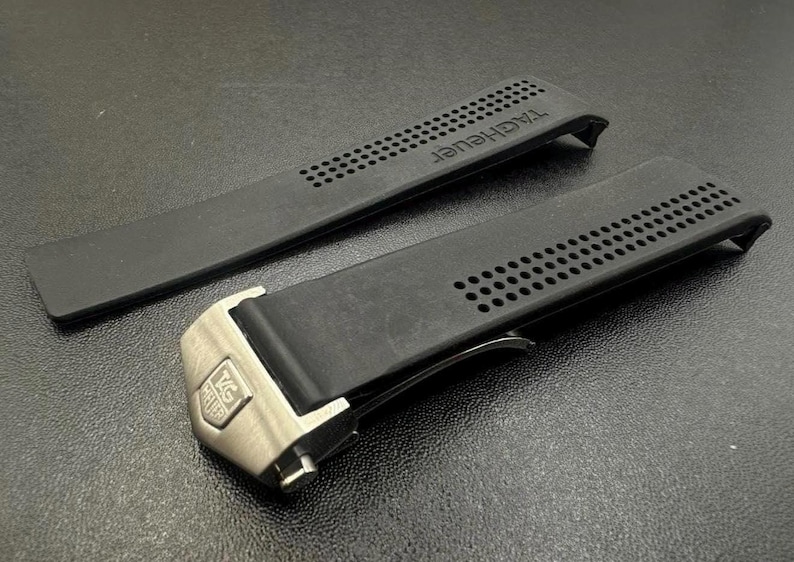 Pu&ograve; includere: Cinturino nero per orologio con il marchio "TAG Heuer" in rilievo. Il cinturino presenta una serie di piccoli fori e una chiusura color argento. Il cinturino &egrave; realizzato in materiale flessibile.
