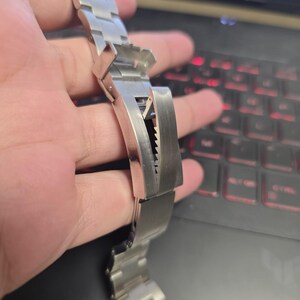 Puede incluir: Una correa de reloj de acero inoxidable con cierre desplegable. La correa está compuesta por múltiples eslabones rectangulares, y el cierre presenta un mecanismo de bloqueo seguro. La correa del reloj se sostiene en una mano.