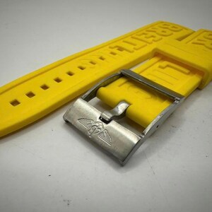 May include: Bright yellow watch strap with the brand name embossed. The strap has a series of square holes for adjustment and is paired with a silver-colored metal buckle. The buckle features a winged emblem.