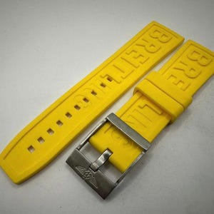 May include: Bright yellow Breitling watch strap with a silver-tone buckle. The strap is made of a flexible material and features the brand name embossed on the band. The buckle has the Breitling logo. This watch accessory is designed for a Breitling watch.