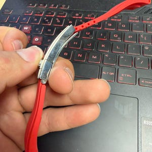 Peut inclure: Un bracelet de montre rouge vif avec un fermoir argent&eacute; est pr&eacute;sent&eacute; sur un clavier d'ordinateur portable. Le bracelet, en mat&eacute;riau souple, comporte des trous de r&eacute;glage. Le clavier a des touches r&eacute;tro&eacute;clair&eacute;es rouges.