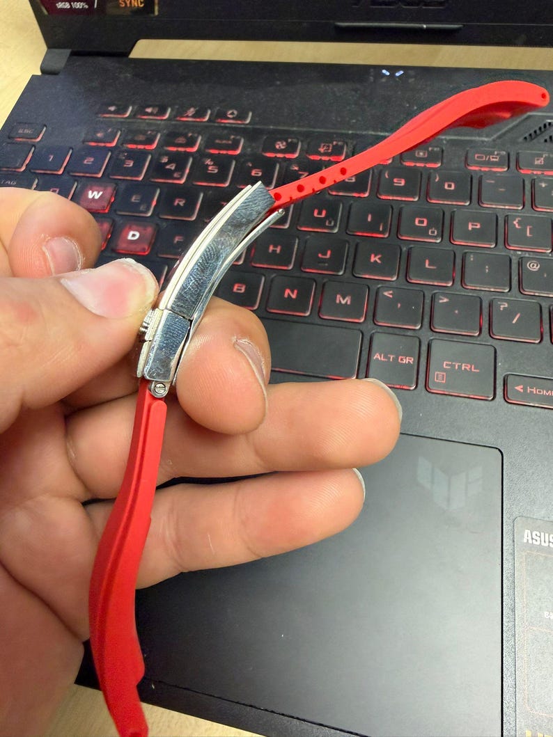 Peut inclure: Un bracelet de montre rouge vif avec un fermoir argent&eacute; est pr&eacute;sent&eacute; sur un clavier d'ordinateur portable. Le bracelet, en mat&eacute;riau souple, comporte des trous de r&eacute;glage. Le clavier a des touches r&eacute;tro&eacute;clair&eacute;es rouges.
