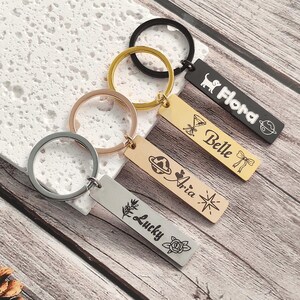 May include: Four rectangular metal keychains with circular rings. The black keychain features the name Flora with a dog and planet icon. The gold keychain displays the name Belle with a glass and bow icon. The rose gold keychain shows the name Aria with a planet and star icon. The silver keychain displays the name Lucky with a branch and rose icon.