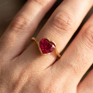 Pode incluir: Um anel de ouro com uma única pedra preciosa vermelha profunda em forma de coração, engastada numa banda de metal liso e polido. O engaste segue o contorno do coração, criando uma linha fluida e contínua que envolve o dedo com um design simples.
