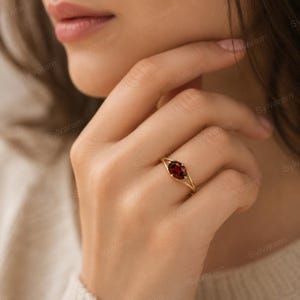 May include: A gold ring featuring a single oval-cut dark red garnet gemstone set in a delicate four-prong setting. The band is thin and polished, showcasing a minimalist design that highlights the deep crimson color of the central stone.