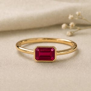 May include: A gold ring featuring a rectangular, emerald-cut deep red gemstone set in a bezel setting. The band is thin, smooth, and polished, showcasing a minimalist design that highlights the vibrant colour and geometric shape of the centre stone.