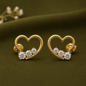 May include: A pair of gold heart-shaped stud earrings, each featuring a smooth, polished gold frame in the outline of a heart. Along the lower curve of each heart, three round, clear, faceted stones are set in a row, adding sparkle to the design.