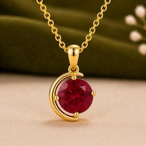 Peut inclure: Un collier en or avec un pendentif circulaire orné d'une pierre précieuse rouge vive insérée dans un cadre en forme de croissant de lune. Le pendentif est suspendu à une fine chaîne dorée, avec une finition polie et une pierre ronde facettée.