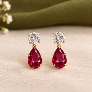 Peut inclure: Une paire de boucles d'oreilles pendantes en or avec une pierre ronde transparente taille brillant en haut et une pierre rouge foncé en forme de poire suspendue en dessous. Chaque boucle est fixée par une monture en or maintenant les deux pierres.