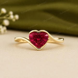 May include: A gold ring featuring a heart-shaped, deep red gemstone set in a smooth, polished metal band. The band curves gracefully around the base of the heart, creating a continuous, flowing design that highlights the vibrant red stone.