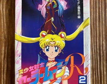 May include: A softcover book titled Sailor Moon R 2 featuring a colorful anime illustration of a blonde character with long pigtails in the foreground and two figures with pink and blue hair standing above against a dark blue and pink background.