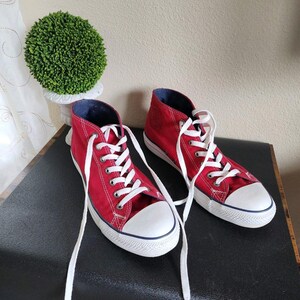 Könnte beinhalten: Ein Paar rote High-Top-Sneaker mit weißen Schnürsenkeln und einer weißen Zehenkappe. Die Sneaker haben ein marineblaues Innenfutter und eine weiße Sohle mit einem marineblauen Streifen. Die Schuhe stehen auf einer schwarzen Oberfläche.