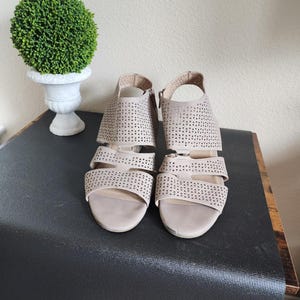 Könnte beinhalten: Ein Paar beige Sandalen mit offener Spitze und Ausschnittdetails. Die Sandalen haben einen Blockabsatz und einen Knöchelriemen. Links von den Sandalen steht eine kleine, runde, grüne Topiary in einer weißen Urne.