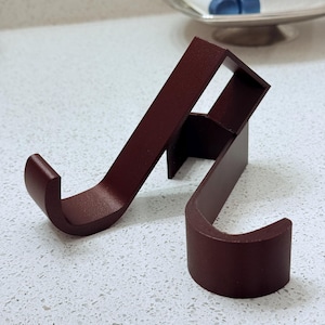 May include: Two dark burgundy metal hooks, each with a curved end, are positioned on a speckled white surface. The hooks are designed to hold items and have a minimalist, modern aesthetic.