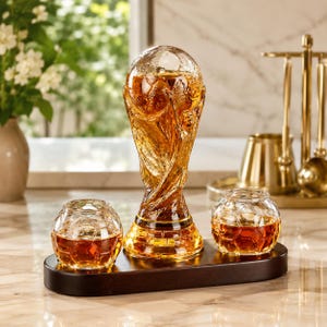Puede incluir: Un juego de tres vasos de cristal llenos de líquido ámbar, presentado sobre una bandeja de madera oscura. El vaso central tiene forma de trofeo, flanqueado por dos vasos redondos. Los vasos son probablemente para servir licores.