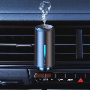 May include: A cylindrical, blue and black car air freshener with a blue light strip. The air freshener is emitting a wisp of vapor. It is mounted in a car's air vent, with the dashboard and controls visible below.
