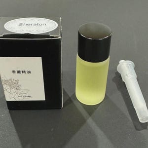 Puede incluir: Una pequeña botella de vidrio con una tapa negra y un aplicador de plástico transparente junto a una caja negra. La botella contiene un líquido amarillo. La caja tiene la palabra "Sheraton" y texto en chino.