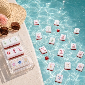 May include: White and red Mahjong tiles with various symbols are displayed in a clear acrylic tray. Several tiles and red dice float in a pool of blue water. A straw hat and sunglasses sit on the pool's edge, suggesting leisure.