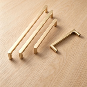 Puede incluir: Cuatro tiradores de armario de metal dorado con un acabado mate liso. Tres de los tiradores son largos y delgados con un perfil rectangular, mientras que el cuarto tirador es más corto y está colocado horizontalmente a la derecha.