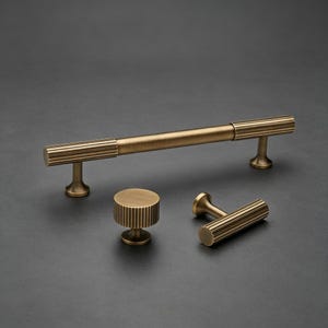May include: A set of four brass-toned cabinet hardware pieces. The set includes a long bar pull, a round knob, and two smaller pulls. Each piece features a ribbed design on the cylindrical ends and a smooth, matte finish.