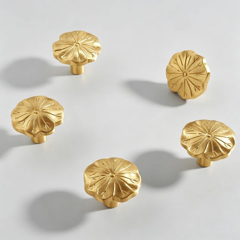 K&ouml;nnte beinhalten: Sechs goldfarbene Schrankkn&ouml;pfe mit floralem Design. Jeder Knopf hat ein detailliertes Bl&uuml;tenblattmuster und einen kurzen, zylindrischen Sockel. Die Kn&ouml;pfe sind auf einer wei&szlig;en Oberfl&auml;che angeordnet und unterstreichen ihre dekorative Wirkung f&uuml;r die Inneneinrichtung.