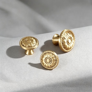 Puede incluir: Tres pomos redondos de latón para armarios con acabado en oro cepillado. Cada pomo presenta un detalle circular central rodeado por un patrón de pétalos y un borde con textura de cuerda. Los pomos tienen una base cónica y una superficie plana.