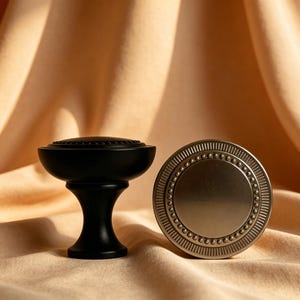 May include: Two decorative cabinet knobs. One is black with a rounded top and pedestal base. The other is a silver-toned, circular knob with a detailed, patterned edge and a smooth center. Both are set against a neutral fabric backdrop.