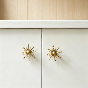 May include: Two gold-colored sun-shaped cabinet knobs are mounted on a white cabinet. The knobs have a central circular design with radiating wavy lines. The cabinet doors are closed, and the background is a light wood.