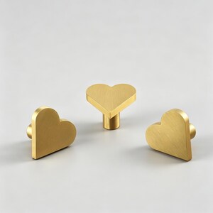 May include: Three heart-shaped cabinet knobs in a brushed gold finish. One knob is upright, while the other two are positioned on their sides. The knobs have a simple, modern design, suitable for drawers or cabinets.