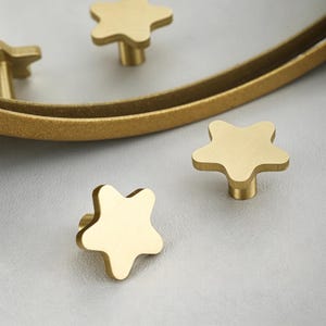 May include: Gold-colored star-shaped cabinet knobs. The knobs have a brushed finish and a short, cylindrical base. Several knobs are visible, along with a curved, gold-colored metal accent piece, all set against a light gray background.
