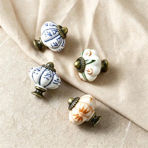 Peut inclure: Quatre boutons de placard en céramique avec une forme arrondie et cannelée et des bases en métal couleur laiton antique. Deux boutons présentent des motifs peints en bleu sur fond blanc, tandis que les deux autres affichent des motifs floraux orange et verts sur blanc.