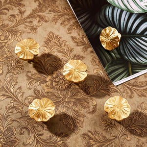 K&ouml;nnte beinhalten: F&uuml;nf goldfarbene Schrankkn&ouml;pfe mit einem Blumenmuster sind auf einer braunen Oberfl&auml;che mit floralem Muster angeordnet. Die Kn&ouml;pfe haben ein detailliertes Bl&uuml;tenblattdesign und bestehen aus gl&auml;nzendem Metall. Der Hintergrund enth&auml;lt eine Zeitschrift mit einem gr&uuml;nen Blatt.