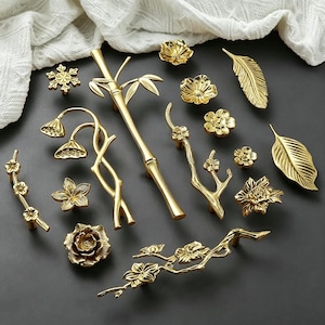 May include: A collection of gold-colored cabinet hardware, including knobs, pulls, and handles. The designs feature floral, leaf, and bamboo motifs. The hardware is arranged on a dark gray surface, with a white fabric in the background.