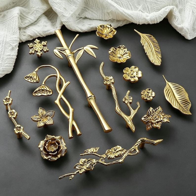 May include: A collection of gold-colored cabinet hardware, including knobs, pulls, and handles. The designs feature floral, leaf, and bamboo motifs. The hardware is arranged on a dark gray surface, with a white fabric in the background.