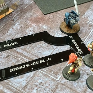 May include: A black game board tool with white text indicating "3" PILE IN", "6" MOVE", and "8" DEEP STRIKE". Miniature figurines with orange hair and swords are positioned near the tool. A blue figurine is also present.