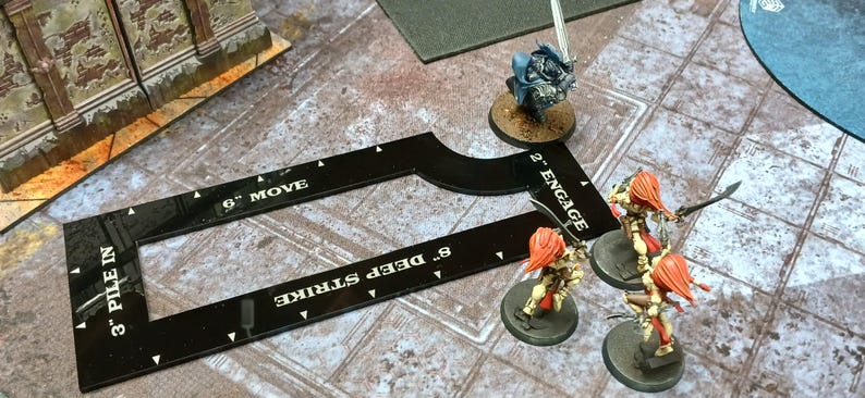 May include: A black game board tool with white text indicating "3" PILE IN", "6" MOVE", and "8" DEEP STRIKE". Miniature figurines with orange hair and swords are positioned near the tool. A blue figurine is also present.