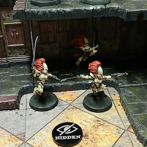 May include: Miniature figurines of female warriors with red hair and armour, wielding swords and guns, are posed on a detailed game board. A black token with the word "HIDDEN" is also present.