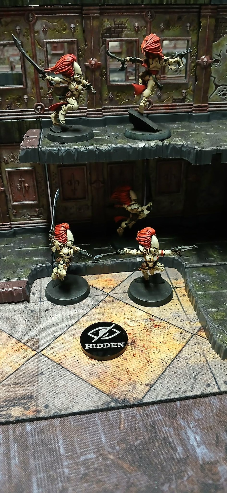 May include: Miniature figurines of female warriors with red hair and armour, wielding swords and guns, are posed on a detailed game board. A black token with the word "HIDDEN" is also present.