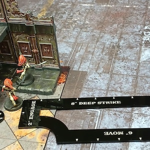 May include: A wargaming scene with miniature figures and terrain. Figures with red hair and armour are positioned near a building. A black measuring tool with text is in the foreground. The scene is set on a textured, grey surface.