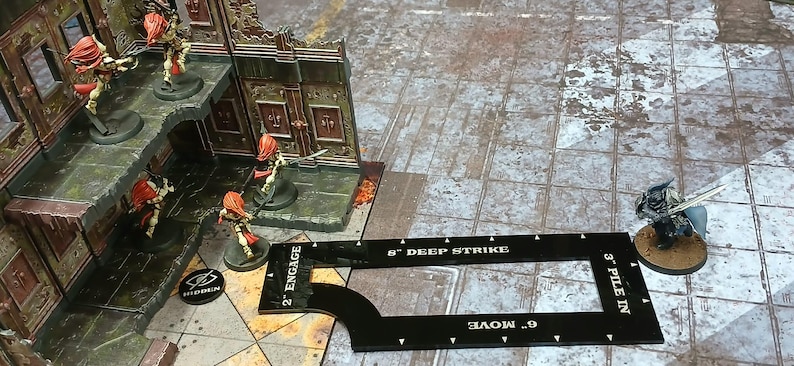 May include: A wargaming scene with miniature figures and terrain. Figures with red hair and armour are positioned near a building. A black measuring tool with text is in the foreground. The scene is set on a textured, grey surface.