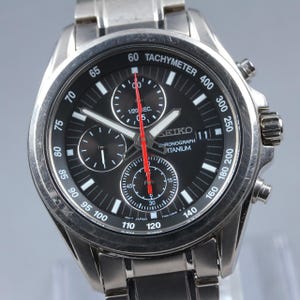 May include: A silver-toned Seiko chronograph watch with a black dial, featuring a tachymeter scale on the bezel, three sub-dials, a date window at the three o'clock position, and a bright red central second hand, all crafted from titanium.