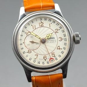 Könnte beinhalten: Eine automatische Oris-Armbanduhr mit einem silberfarbenen Edelstahlgehäuse und einem strukturierten cremefarbenen Zifferblatt. Die Uhr verfügt über arabische Ziffern, ein Hilfszifferblatt, einen Datumszeiger mit roter Mondsichelspitze und ein orangefarbenes Lederarmband.