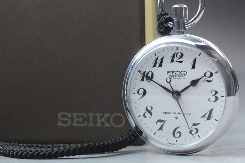 以下が含まれることがあります： 白文字盤と黒数字のシルバーSeiko Precision懐中時計。時計には黒いコードが付いており、Seikoのロゴが入った濃い灰色の箱の隣にあります。文字盤には「SEIKO PRECISION 17 JEWELS SECOND SETTING」と書かれています。
