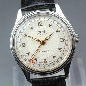 May include: A vintage Oris automatic wristwatch with a silver-tone case and a black leather strap. The watch face is off-white with gold-tone hands and markers. The dial includes the text "ORIS 17 JEWELS AUTOMATIC". The watch also displays the date.