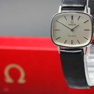 Pode incluir: Um relógio de pulso Omega Genève em tom prateado com caixa retangular e cantos arredondados. O relógio apresenta um mostrador claro com marcadores de hora pretos, ponteiros pretos e o logótipo Omega. Possui uma bracelete de pele preta lisa.