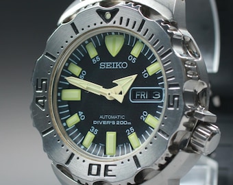 May include: A stainless steel Seiko automatic diver's watch with a black dial. The watch features luminous hands and markers, a date display, and a black rubber strap. The bezel has a rotating design with minute markers.