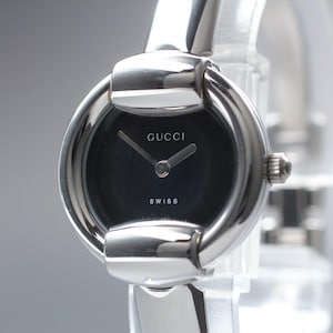 May include: A silver-toned Gucci watch with a black face. The watch features the brand name "GUCCI" and the word "SWISS" on the face. The watch has a round face and a silver band.